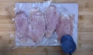Flattened chicken breasts covered in plastic wrap being tenderized on a cutting board.